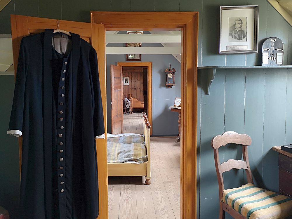 This photo looks through a doorway, through a room, and into a third room. In the nearest room, the walls are wood and painted a grayish blue. A pastor's robe hangs on the back of the open door on the left. On the right is a chair at a desk. The light comes from a window (out of frame) above the chair. On the wall above the chair and next to the door is a fraimed portrait. IN the next room the end of a double bed is visible. On the wall next to the next door is a clock. Through that door the end of a single bed is visible.