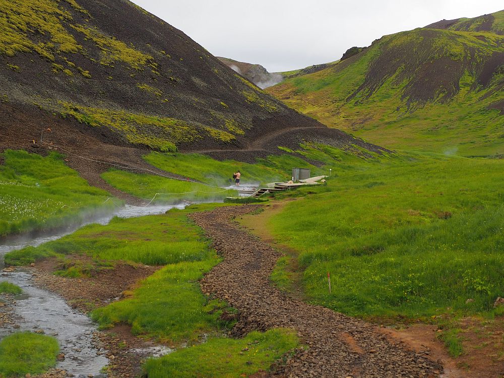 The path stretches ahead in the middle of the picture, down a green valley with mountains on both sides. The river flows diagonaly across the image from lower left to upper right. In the center of the photo is a boardwalk along the river, with a wall at one point (described in the article) and a couple of people are visible too. 