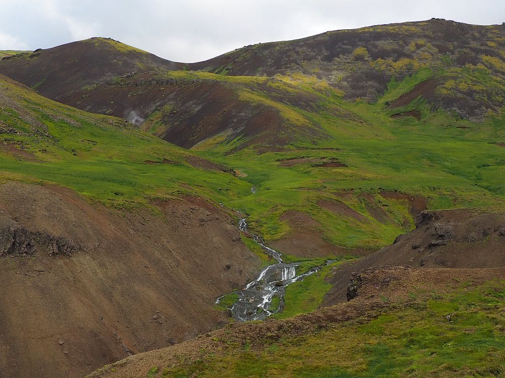 Mountains in the background, and, in the foreground, a waterfall flows down a narrow green valley in a number of steps. 