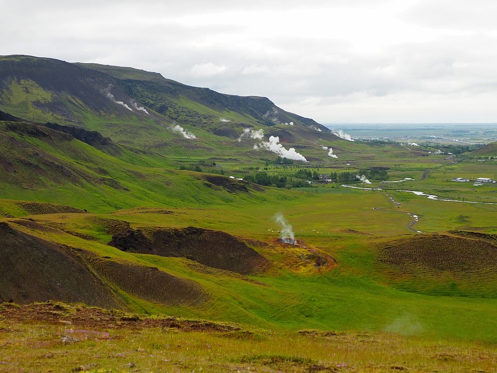 A view down a green valley with a low mountain on the left. Here and there plumes of steam rise from the ground. In the distance, the sea is visible.