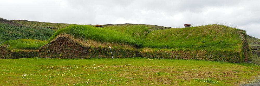 The house is low to the ground. Only the bottom of the walls is brown; the rest is all green with grass, as is the land in front of it. The photo shows two wings of the house, each long and with a simple peaked roof and no windows.