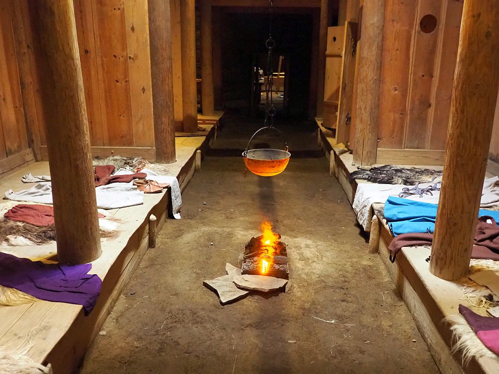 A view down the center of the Viking house's interior. Down the center: a wide dirt floor stretching the length of a long room. On either side, wide low benches about the width of a double bed. Large poles (tree trunks) extend upwards from the benches to the ceiling. In the center of the floor is a small (fake) fire with an iron bowl hanging above it.