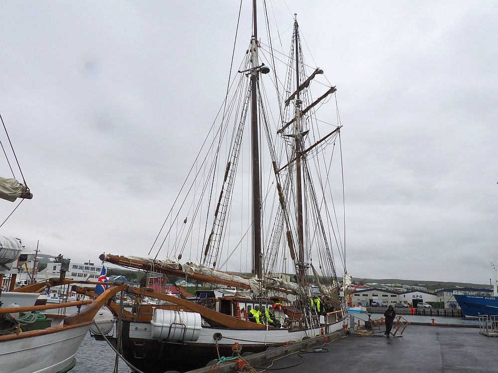 The ship is long and narrow, painted white around the sides but darker further down. It is moored along a pier. It has two masts, each with a web of rigging but the sails are all furled along the spars and booms. Grey cloudy sky behind.