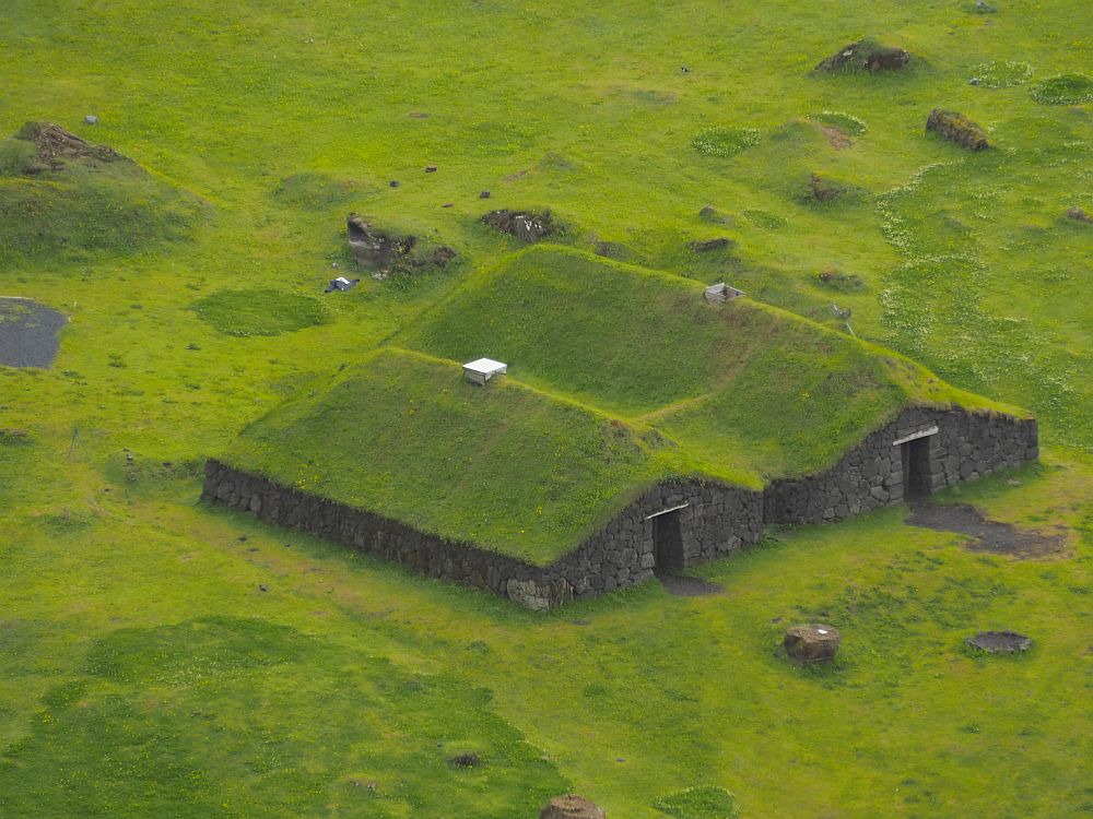 Seen from an angle above, the roofs are green and the field around the building is also green. It has two long rooms, each with a peaked roof. The holes in the center of each roof are capped with something that may be modern metal or plastic. The walls are quite low and built of stone. They The end of both halves of the building has a door, and the building is high enough in the middle for a person to stand upright. At the sides, though, these walls are very low, about half as high as the center under the peaks.