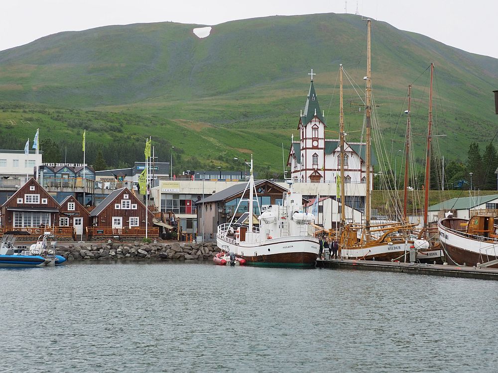 Husavik whale watching leaves from this harbor. Cluster of buildings along the water's edge, closer up than in the image above. Several large oak boats moored along a pier on the right. A white church with a tall tower behind the boats. On the left, a row of 3 red wooden houses with white-framed windows line the waterfront.