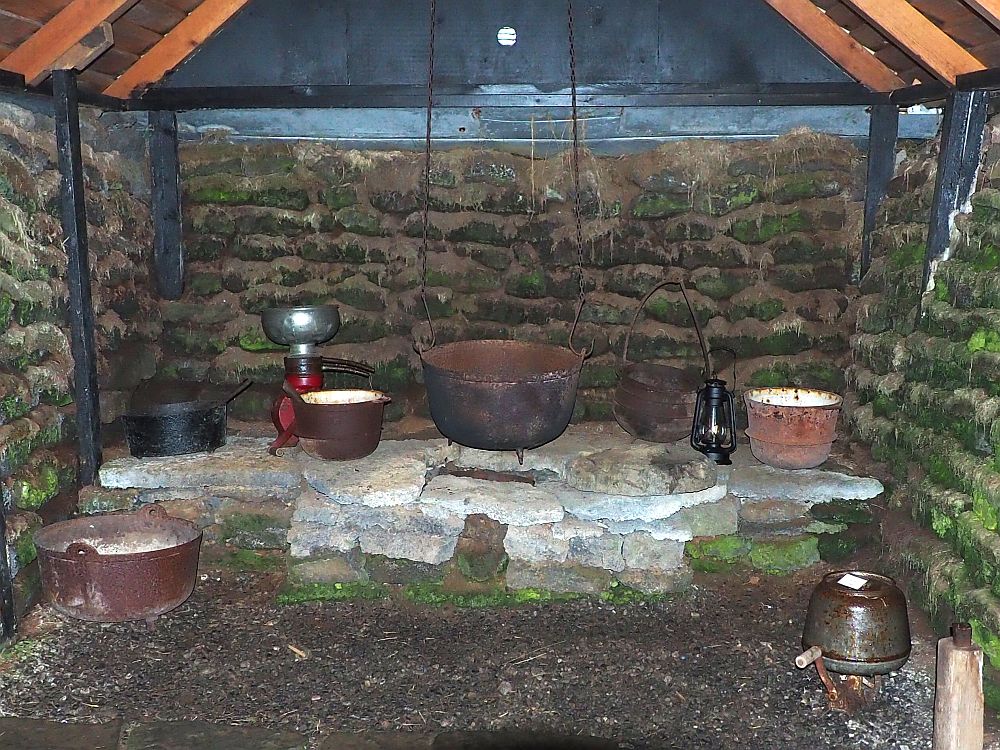 The "stove" is a pile of flat stones built into the end of the room. On top are several iron pots, and one large pot hangs on chains from the ceiling. The walls behind the stove and on either side are turf, with green moss growing on it in some places. Beams of wood stand vertically against the walls and hold up the beams that support the ceiling.