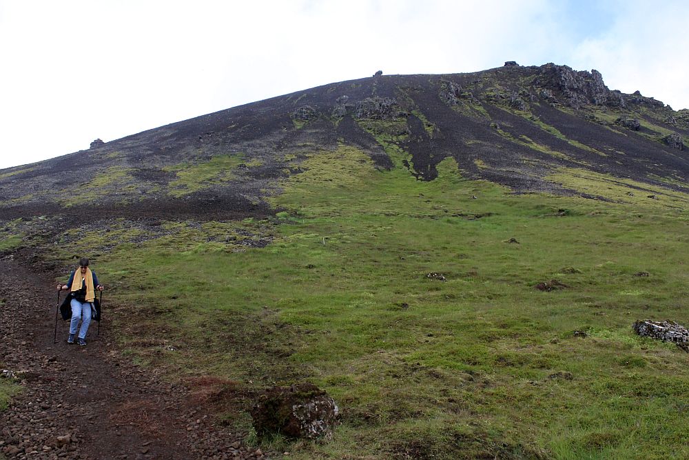 A hill, covered in low grass or moss but with exposed rock on the top. On the left of the picture I am walking down a gravel path, holding two sticks and looking down at the ground. On my way to Reykjadalur hot spring thermal river.