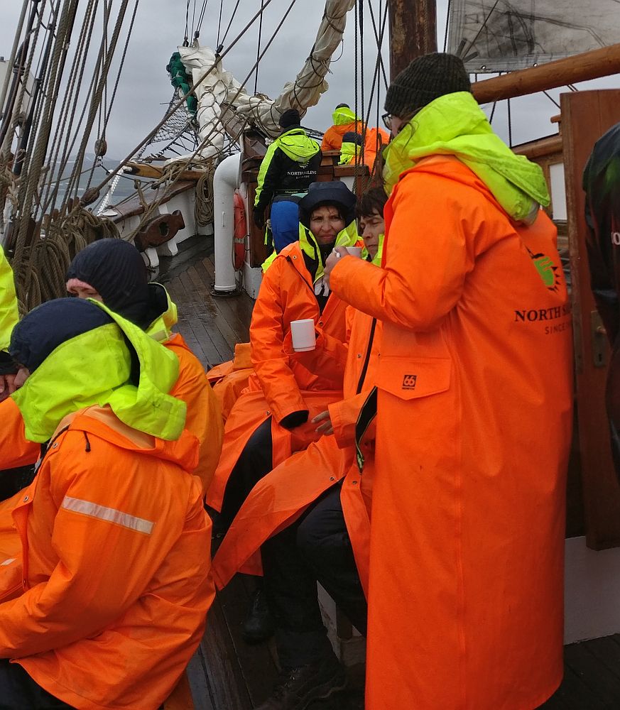 A group of people sit in two rows, each wearing bright orange raincoats. The yellow collars of their coveralls show, sticking out from the necks of their raincoats. Behind them the bow (front) of the ship is visible, rigging with a furled sail.