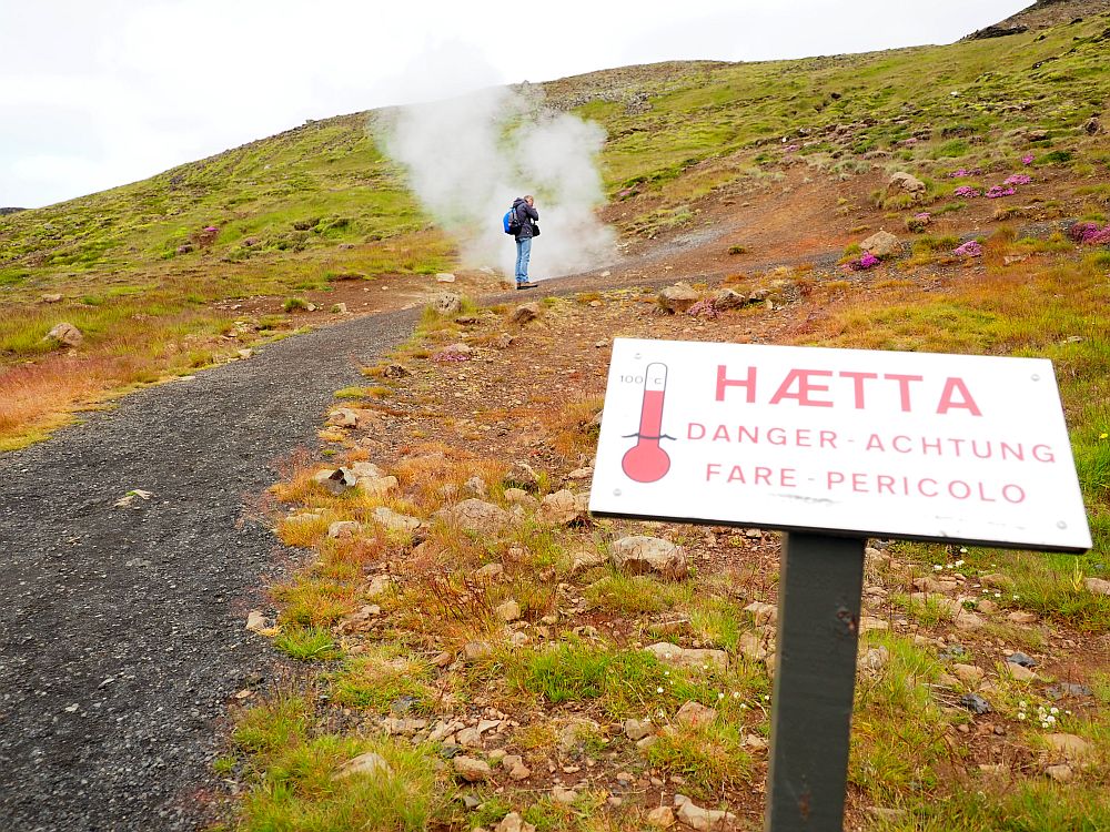 In the foreground a sign with a picture of a thermometer showing a lot of mercury. It says "HAETTA DANGER ACHTUNG FARE PERICOLO". To the left of the sign is a gravel path up a small hill. About halfway up the hill a plume of steam rises from the ground and a man (my husband, Albert) stands right next to it, taking a picture downward into the hole.