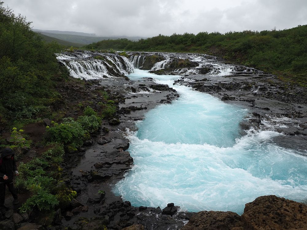 Bruarfoss is seen from a different view here. It is at the center background. In the foreground is the river that flows out of it between two black stone banks. The water is quite a bright blue here.