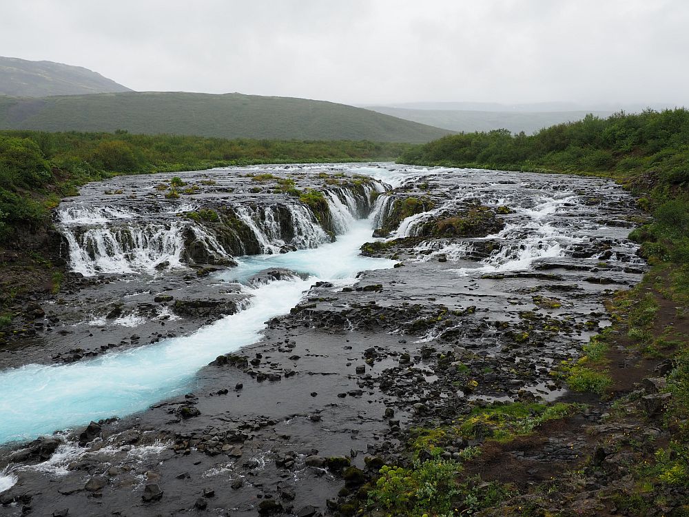 As described in the text, a wide flat place, almost round looking from her, is split by a cleft. Water flows into that cleft from all sides, then into a river that is blueish whte. The rock around it is black, with greenery edging that.