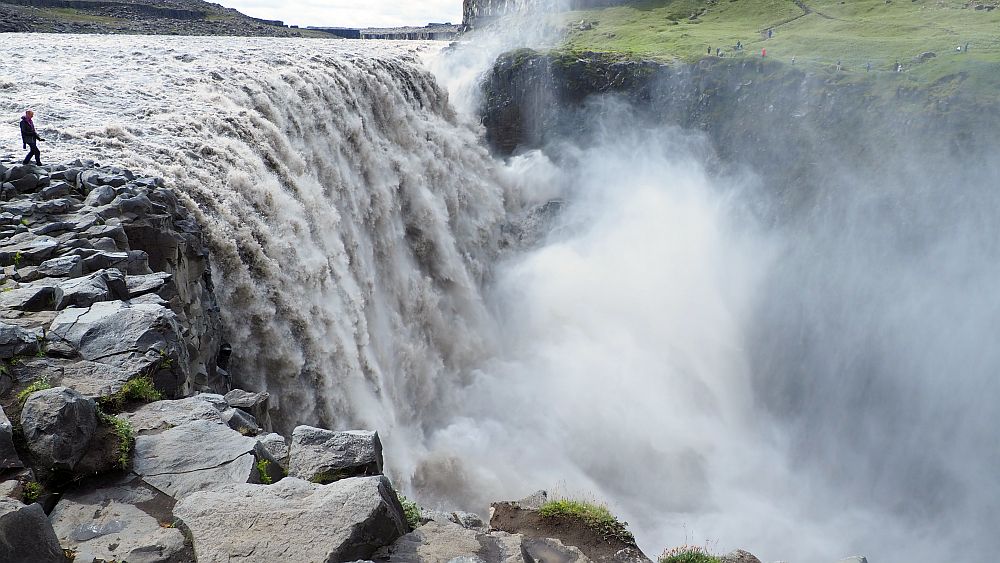 Seen from the level of where the water falls off its cliff shelf, the sheer quantity of water if visible and a lot of spray obscures how far it falls. A person stands right next to the waterfall at the left.