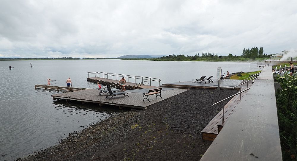 Looking along the lakeshore, the lake on the left. A cloudy sky, so the water looks dark. The beach has quite dark sand. A small pier leads from the beach out onto the lake.