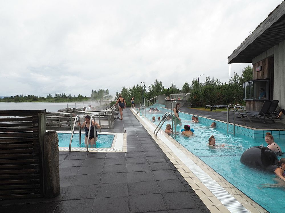 On the right and stretching out straight head is a long shallow pool of clear blue water with a scattering of people sitting in it. On the left, part of another pool is visible. The water is the same color and a woman is climbing the ladder to get in. Steam rises beyond the small pool on the left.