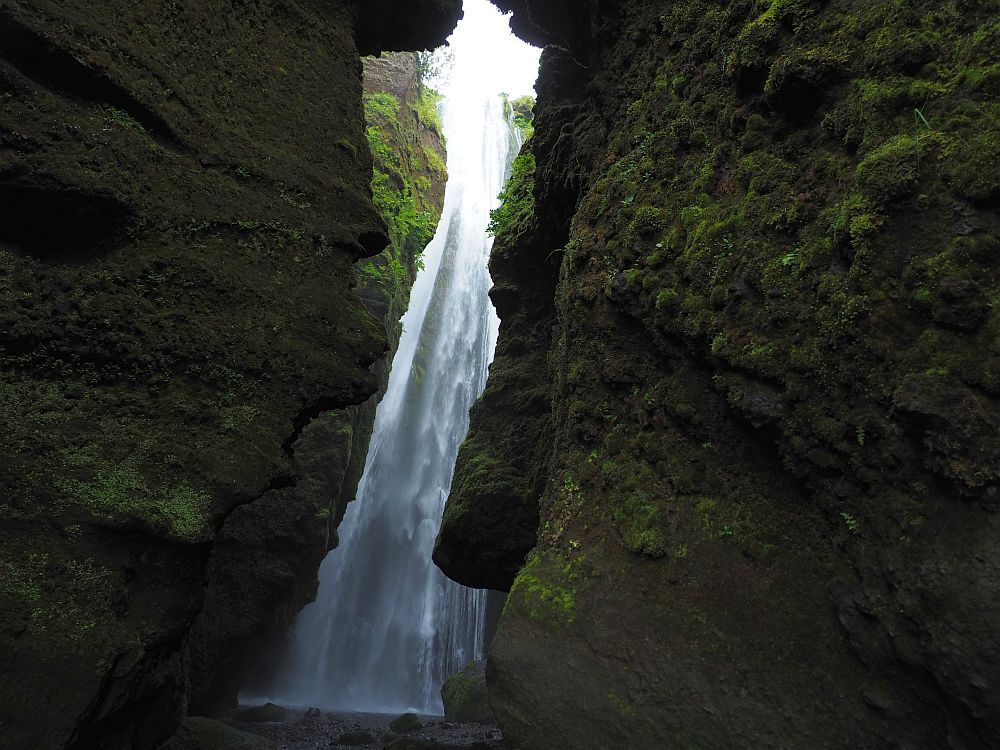 Dark rocky cliffs on both sides of the picture. IN the narrow space between them, the waterfall is visible behind, lit brightly white.