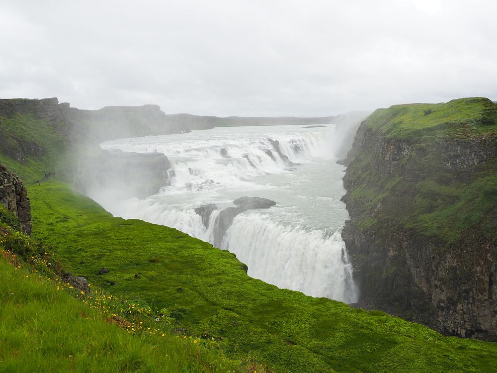 Looking off a cliff to the waterfall in the distance on the opposite side of a canyon. From a distance, the top part of the waterfall looks flat and the water looks all white. It flows toward the camera, then falls off the opposite side of the canyon in a thick white sheet of water.