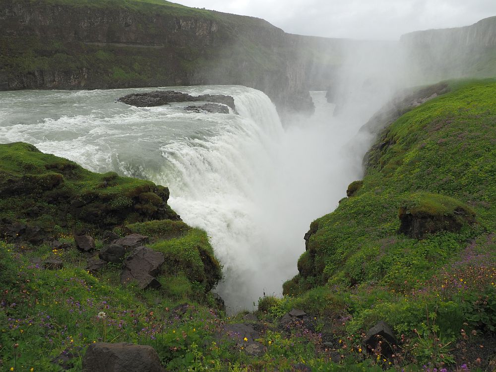 The water flows from the left of the picture and plunges off a wide shelf in the center, into a deep canyon. Clouds of spray obscure the view down the canyon where the water flows next.