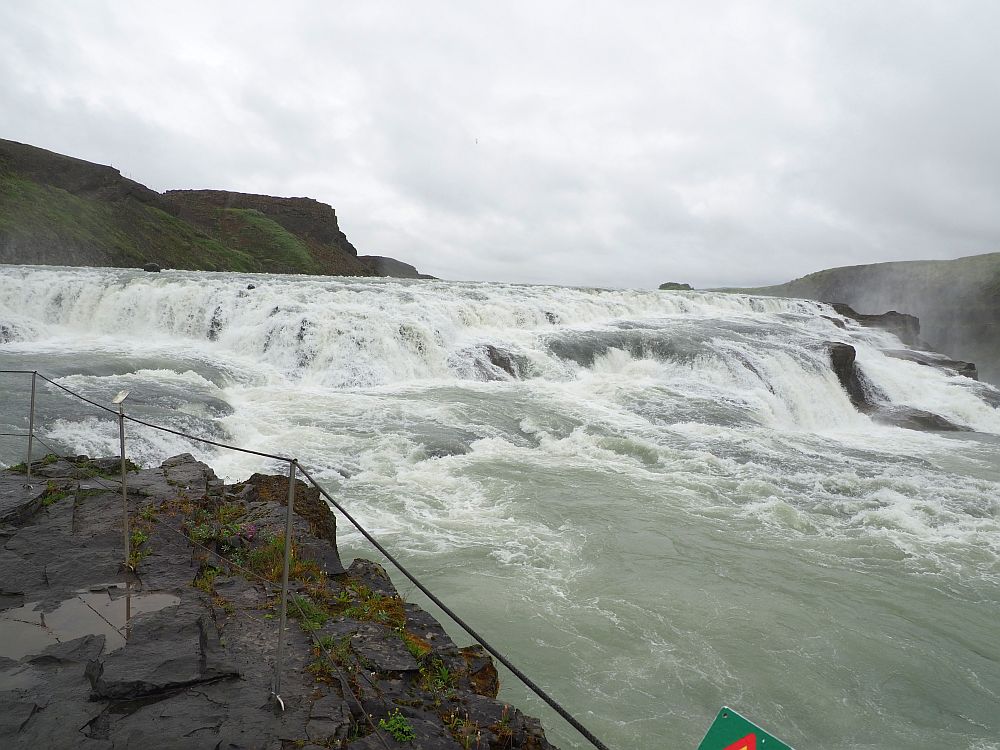 In the foreground, black rock and a rope fencing off the edge. Beyond that, water churning white over a series of rocks.