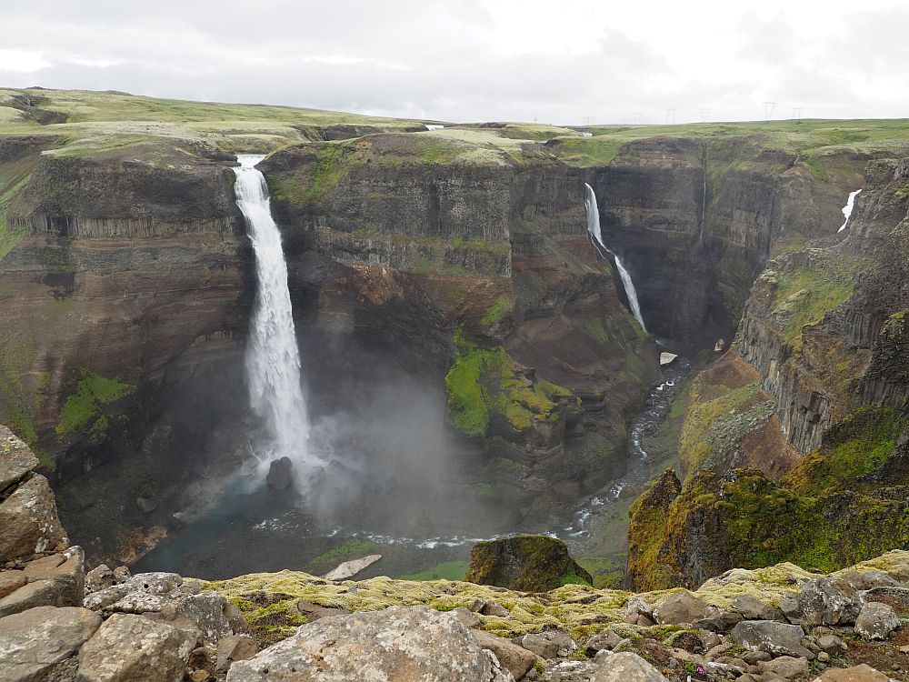 Two waterfalls in this photo: both are long and narrow and fall of the same cliff to a stream in a canyon. Both are white, and the nearer one is wider.