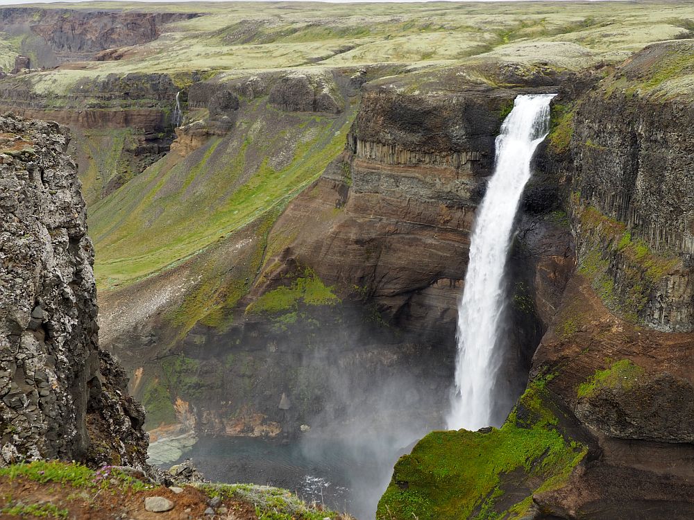 A long narrow white waterfall plunges off a cliff on the opposite side of a deep canyon. The bottom of the falls is not visible. 