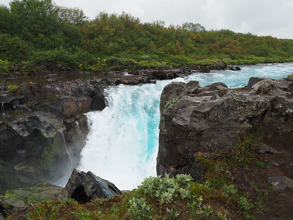 A stream from the top right of the photo flows to the center, then down off a small cliff. Rock on both sides is dark black, with green bushes visible on the opposite side. The water is bright blue, except where it froths white.