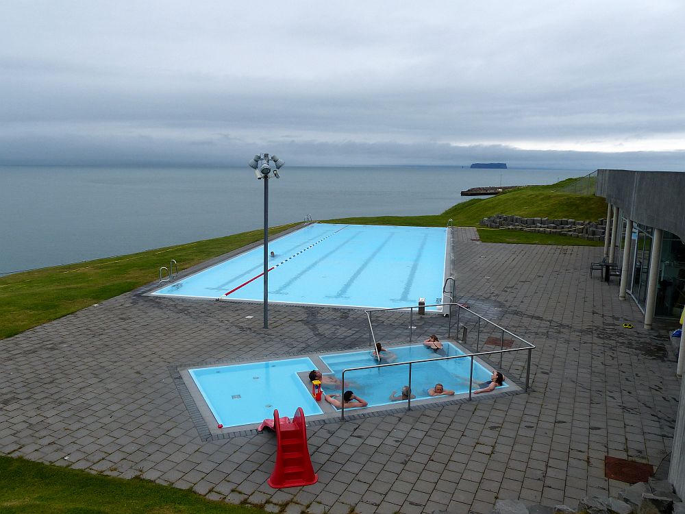 Seen from slightly above, three pools with blue water. Nearest by are two small pools right up against each other: one is a child's wading pool and has a red plastic slide leading into it. The other is a soaking pool and has a number of people sitting on seats along the sides of the pool, soaking. The further pool is more of a normal size, with lanes painted on the bottom and one floating lane divider. Beyond is the sea under a gray sky.