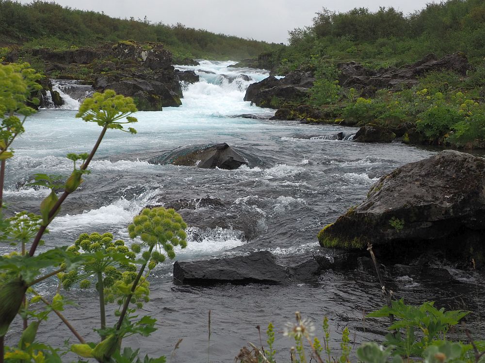 Looking straight upstream, the water flows toward the camera, bumping over various rocks along the way. Rocks on both sides are black, edging the stream, and green bushes are visible on both sides. In places, the water is blue but mostly it is dark grey, except where the rapids look white.