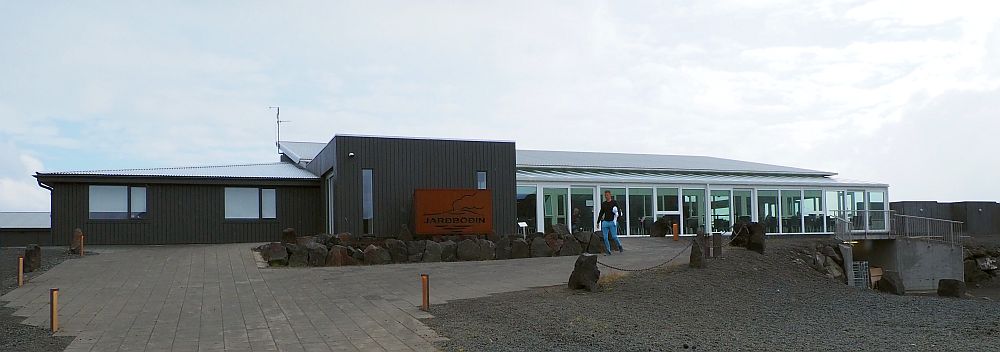 A long low building, brown with a light roof. the left-hand end has just a couple of windows. The right half has a glass wall and tables and chairs are visible inside.