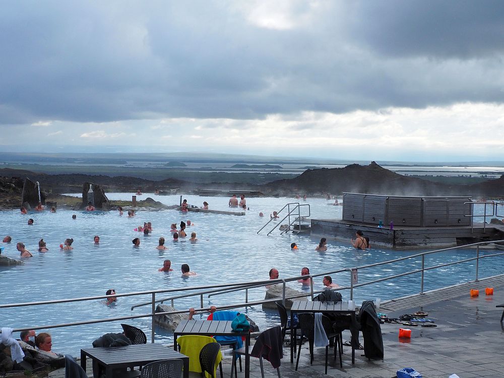 The water looks grayish blue under a gray cloudy sky. There is a railing in front of it along a concrete patio with tables and chairs draped in discarded towels. In the water is a scattering of people and a bit of steam rises from it. Beyond is a flat landscape of brown earth and lakes in the distance. 