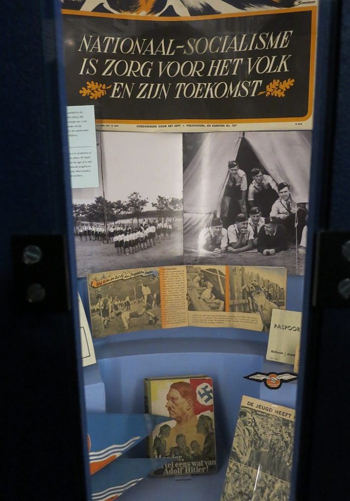 The display is tall and narrow. At the top is the slogan translated in the caption. Under that, photos of Nazi youth lined up in a field on the left and a group of young men in informal uniforms looking out of a tent and smiling on the right. Below that, some publication open to a page of photos of Nazi youth doing activities. On the floor of the display stand several books.