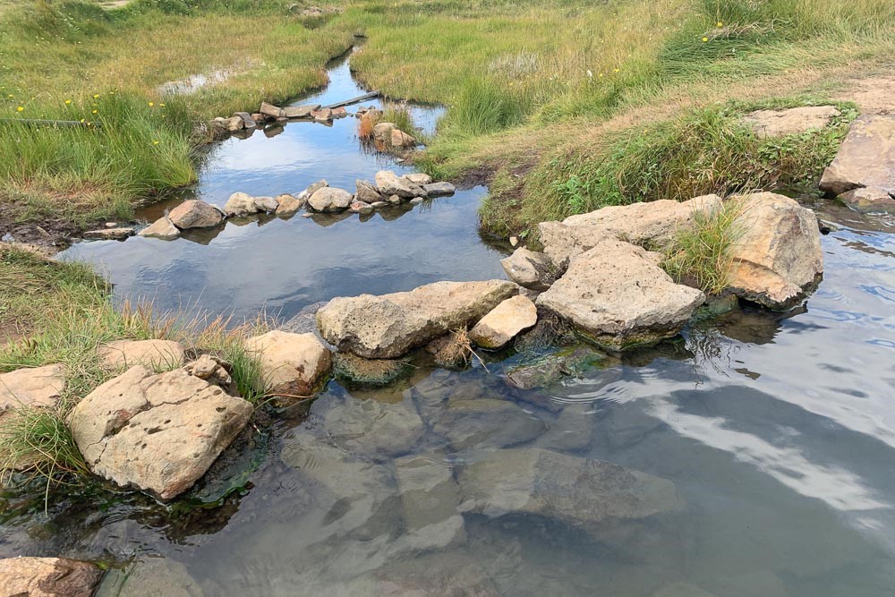 Water in the foreground and a stone wall of sorts across it. Below the stone wall, another pool, then another stone wall and another pool, narrowing to a stream after that. On both sides of the pools and stream, grassy banks.