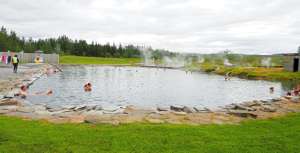 A large pool, edged with large flat stones. A few people are in the water. At the far end clouds of steam rise.