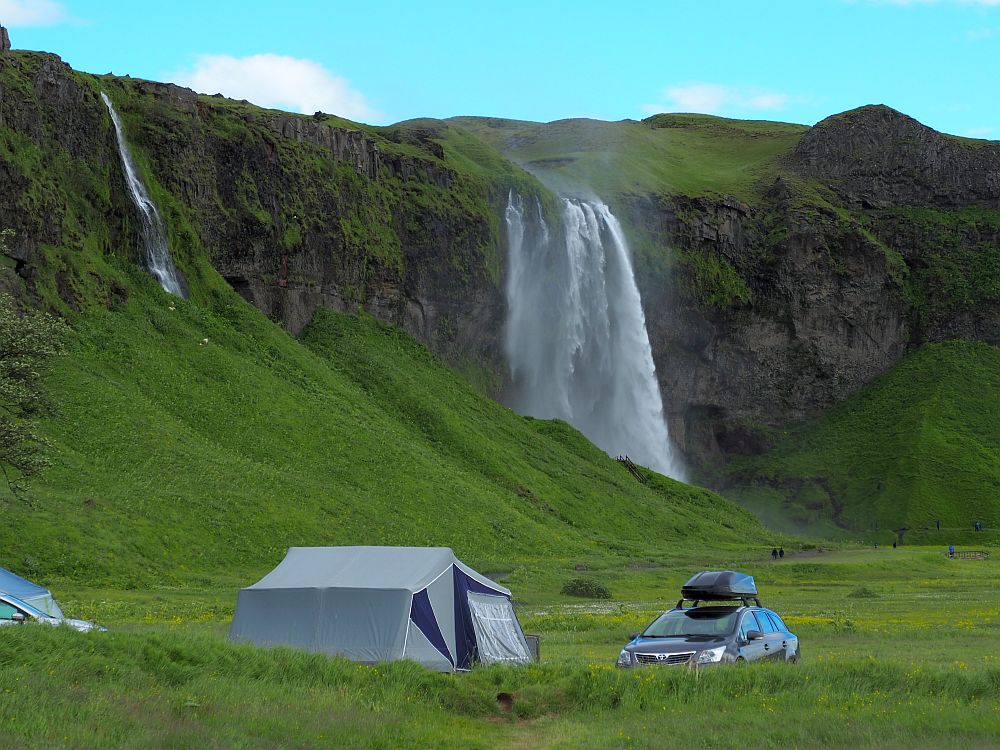 The water falls over a cliff in a veil shape at the back of this picture. In front, at the base of the cliff, is a tent with a car parked next to it. A smaller, narrow waterfall is visible behind the tent to the left.