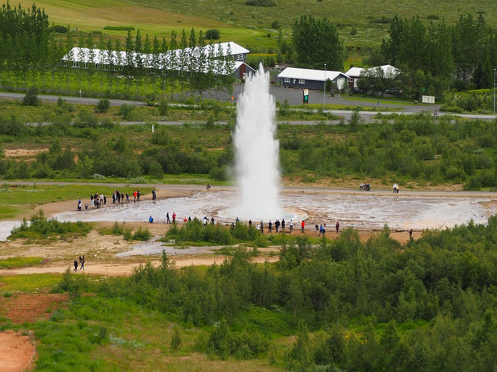 A large flat space as seen from a hill above it. People are visible very small around the edge of the flat area. A huge fountain of white water spouts up in the center of the flat space. Beyond the geyser some low buildings stand across a small street.