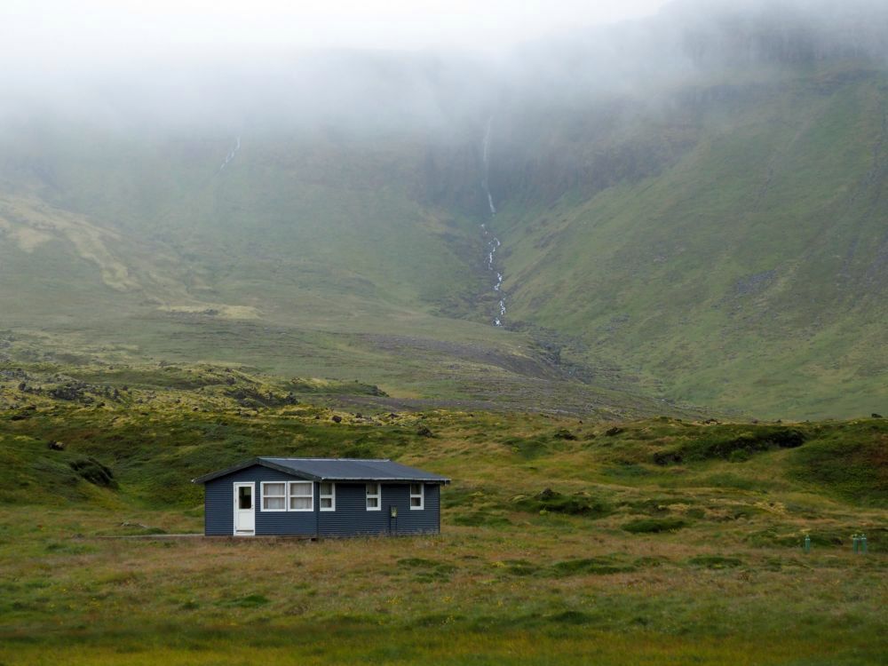 A small grey house with white edging in the foreground. The land behind it is covered in green grass or moss and rises steeply to a mountain, but the top of the picture is obscured by low cloud. In a fold in the mountain a stream is visible, trickling down the mountain.