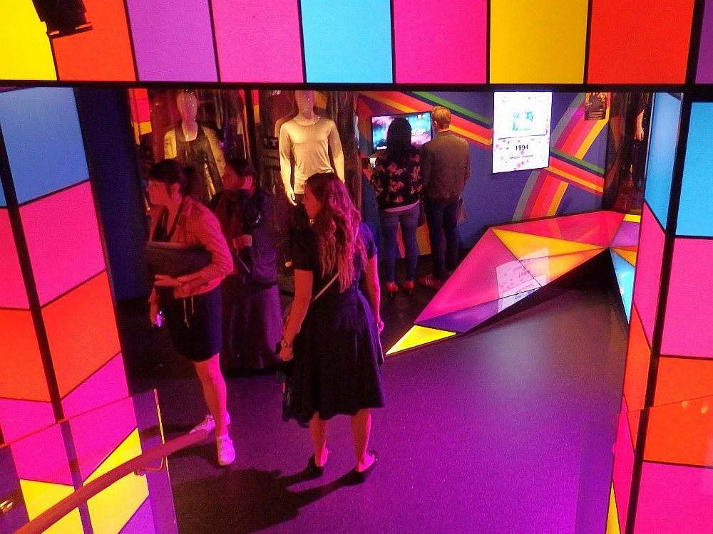 The entrance, looking down at it from upstairs, is framed in brightly-colored cubes of glass, lit from within. Beyond the entrance several visitors are visible.