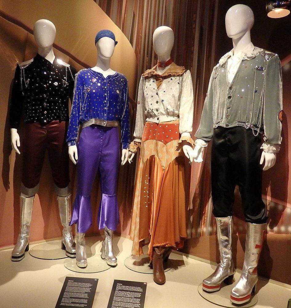 L-R: clothing on mannequins
Bjorn's outfit is black with shiny stars on the shirt and silver-colored shoulder plates. Knee-high boots in silver with platform heels.
Agnetha's outfit is a blue top with silver spots and purple pants with a shiny purple frill above the ankles. Silver platform boots.
Frida's outfit: off-white shirt with multi-colored spots and a brownish collar, orangy-brown flowy skuirt to above the ankles, brown boots.
Bennie's outfit: silver-gray jacket with shiny spots over a white shirt with a big collar, black pants tucked into high silver platform boots.

