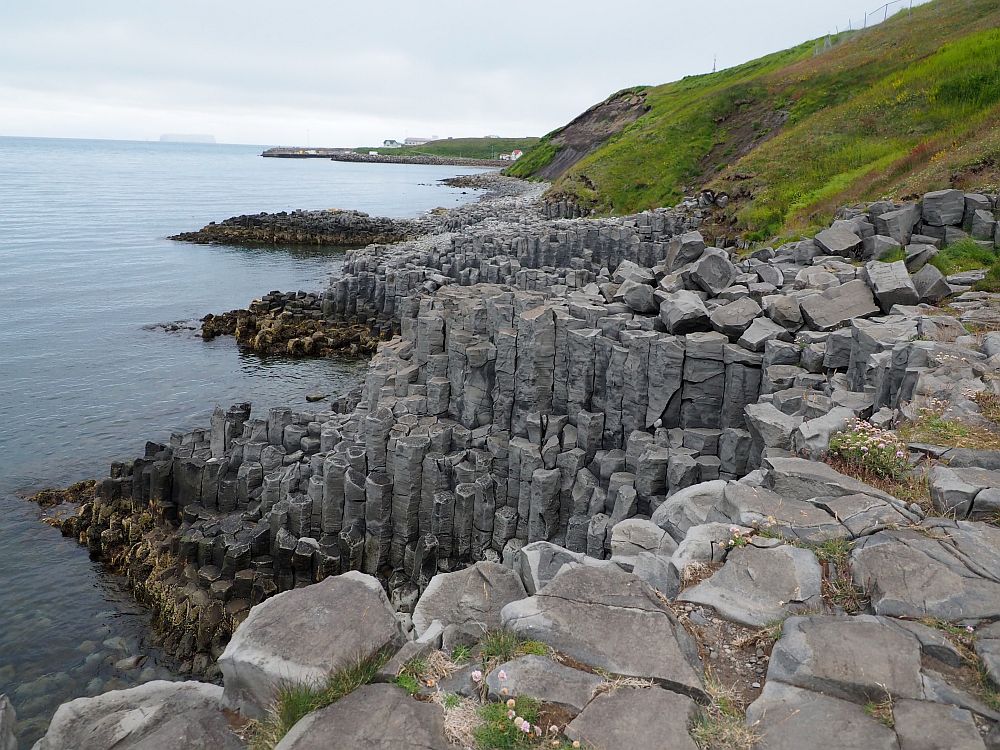 Looking along the coast, sea on the left and moss-covered hill on the right. The coast itself is very rocky, and the rocks are in the form of basalt columns, sticking out in a series of points into the sea.