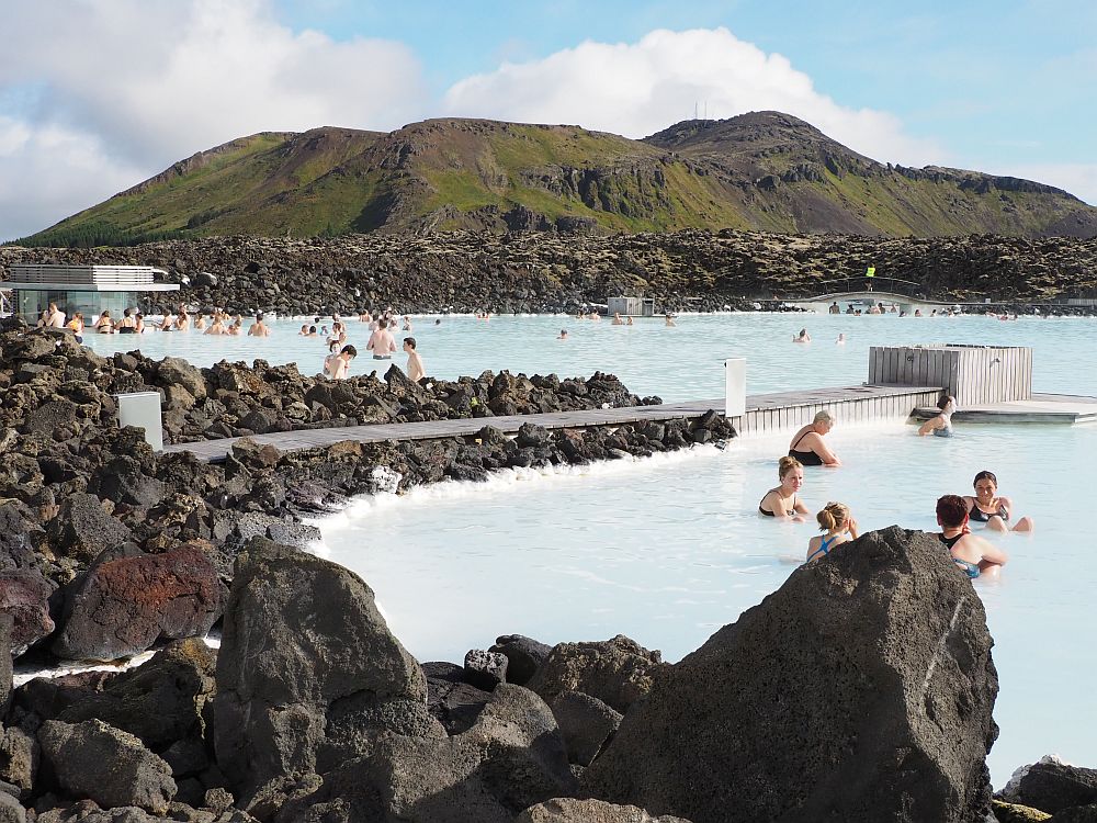 A view of the blue lagoon: milky blue water, edged with piled-up black rocks. People in the water here and there. At the back of the photo is a low hill surrounded by more black rock, covered in spots with green moss.