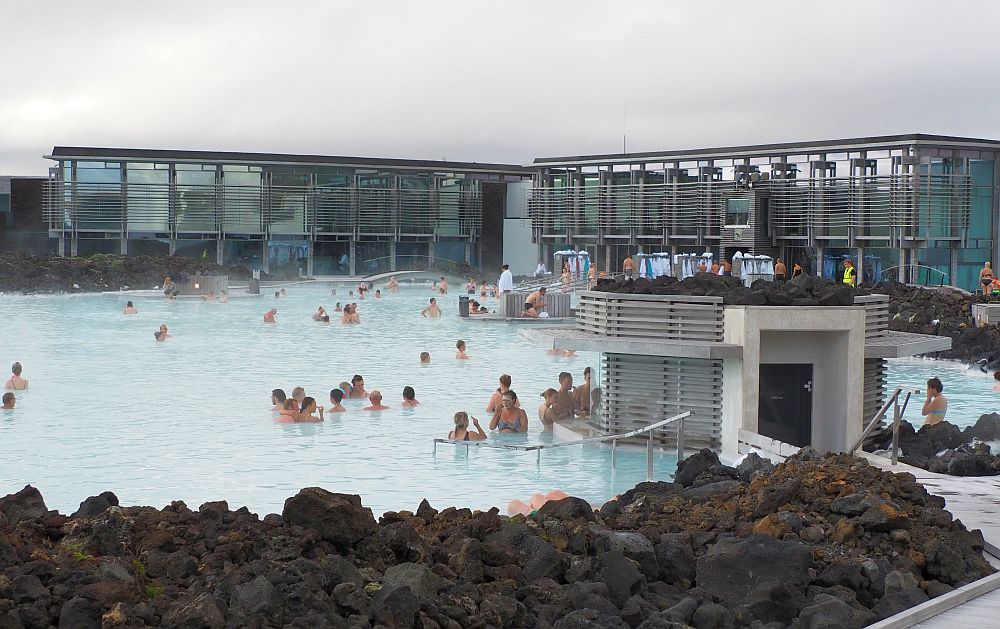 The water of the blue lagoon is milky blue, edged with black volcanic rock. Beyond the water is the modern spa building with lots of glass.