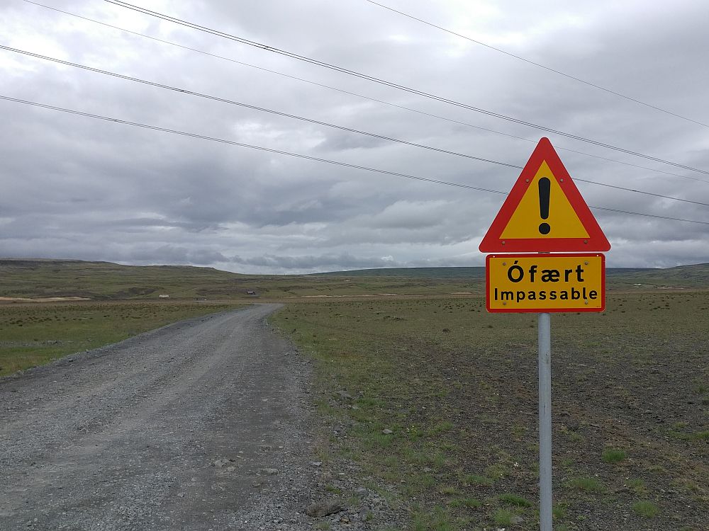 A dirt road extends to the horizon and a sign at its side is a yellow triangle with a red edge and, inside the triangle an exclamation point. Below that, a smaller sign, also yellow with a red edge, but rectangular. It reads "Ofaert Impassable"