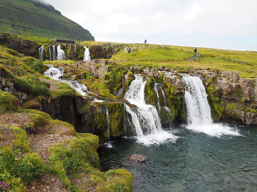 A green hillside, and the white waterfalls move down a series of stone steps, separated into several different streams before the all empty into one pool below.