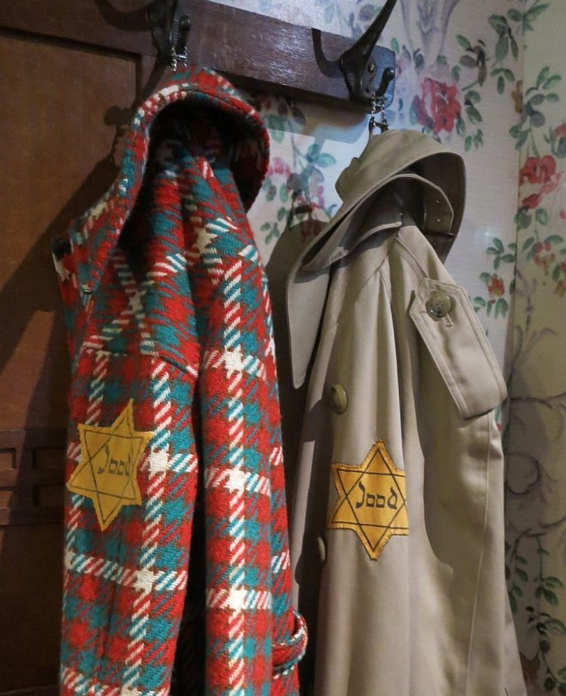 Two coats hang on a coatrack. The left-hand one is brightly checked in red, green and white. The right-hand one is a trenchcoat in beige. Both have a yellow star of David on the sleeve with the word "Jood" inside it, which means "Jew."