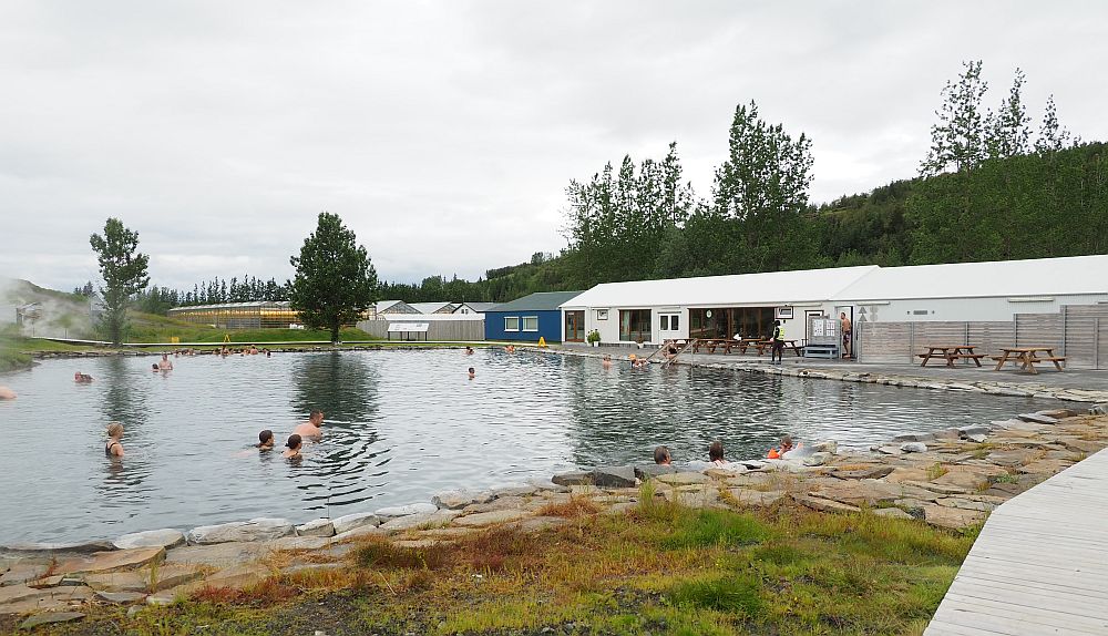 The pool is more or less square, edged with rocks. To the right is a long, low building with a white roof, a few picnic tables in front of it.