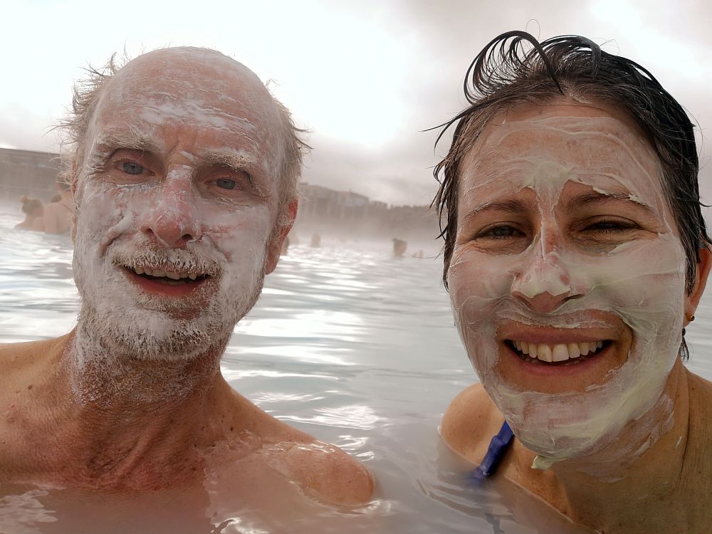 A selfie: my husband on the left, smiling, bald head except some hair on the sides, beard and mustache, face smeared with white mud. On the right, me with my wet hair slicked back, white mud smeared on my face, also smiling. We are up to our shoulders in milky-looking water.