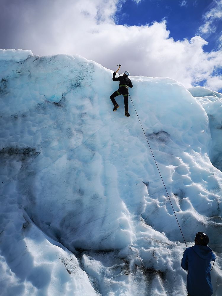 A wall of bluish-white ice. Near the top, a man (Albert), clings to the ice, his feet (crampons) in the ice, about to pound the ice above him with a small pickaxe. He is attached to a rope, which extends down to a person at the bottom of the picture.