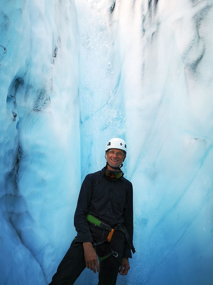 On the Troll Expeditions Iceland glacier hike, Albert stands, smiling at the camera and looking relaxed. He's wearing all black, except for green thing strapped around his waist and loosely between his legs. On his head is a rounded white helmet. Around him are walls of blueish white ice.