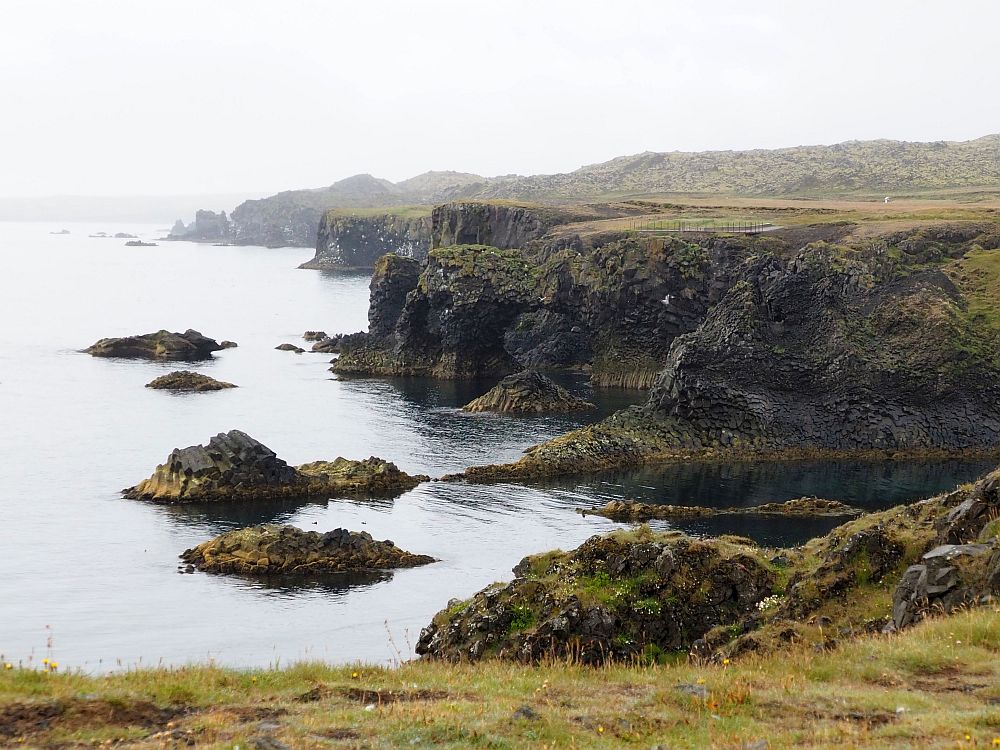 The coastline isn't straight but is lined with cliffs that extend in someplaces outwards, sometimes have little bays between them. Flat on top, steep cliffs down to the water. It's a gray day, so the coast in the background is misty.