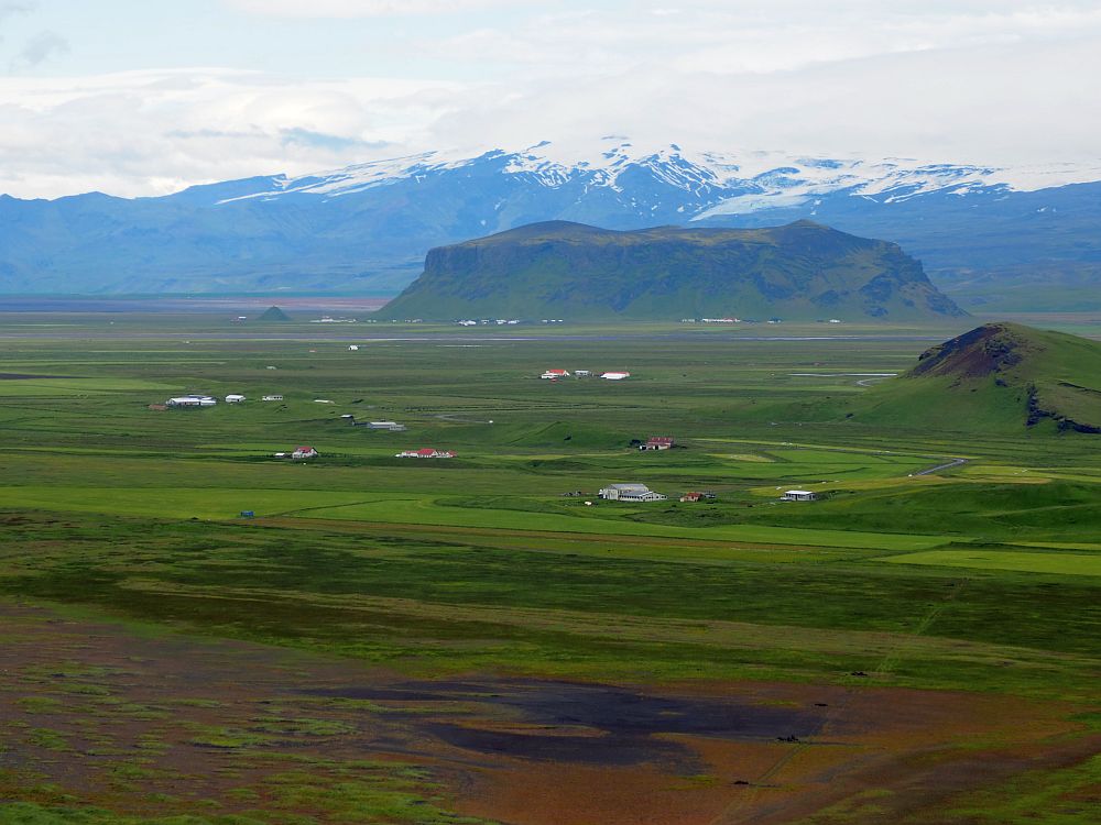 IN the foreground, green fields dotted with the occasional house. Beyond that, another promontory like Dyrholaey. Beyond that, a mountain range, topped with snow.