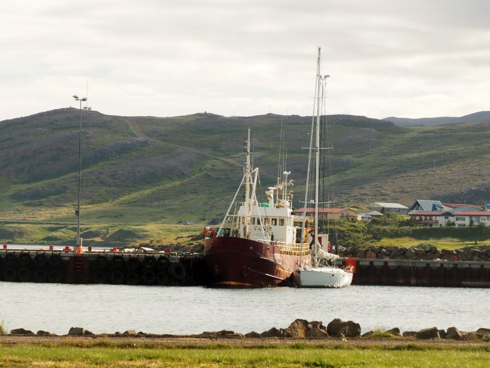 Across a small harbor, two boats moored on the other side. One is a fishing boat, rigged for nets and such, with a reddish hull. Beside it, a much smaller sailing boat with a tall mast.